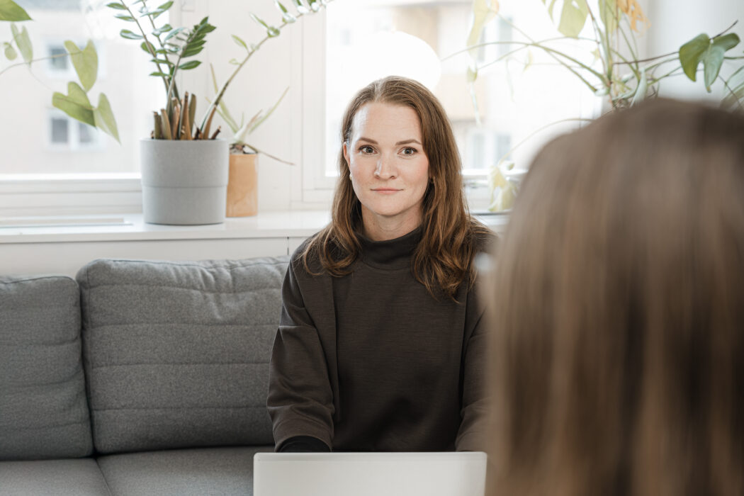 En person sitter på en grå soffa med en laptop framför sig, omgiven av gröna växter i krukor på ett fönsterbräde.