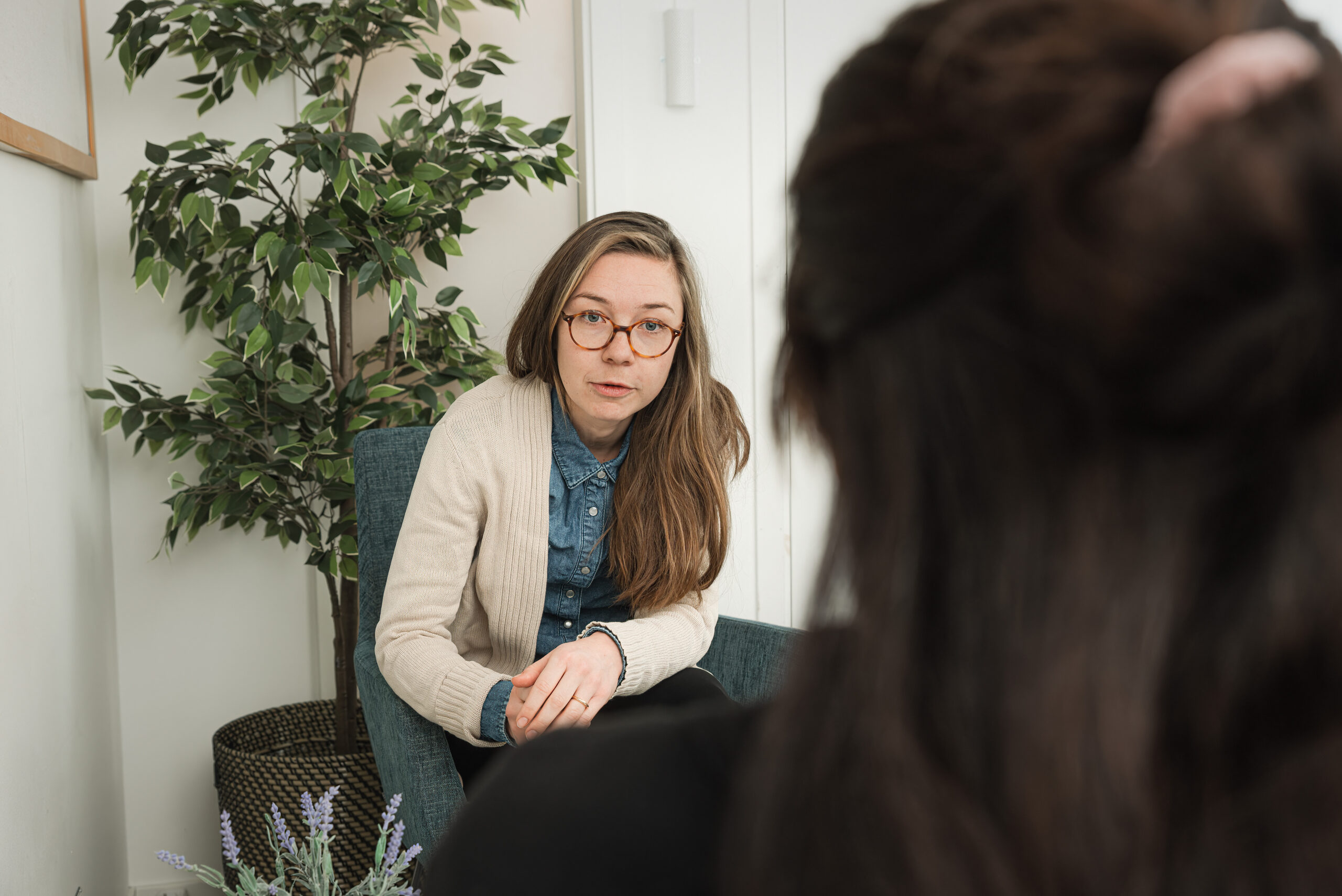 En person sitter i en fåtölj med en grön växt i bakgrunden och en annan person syns delvis i förgrunden.