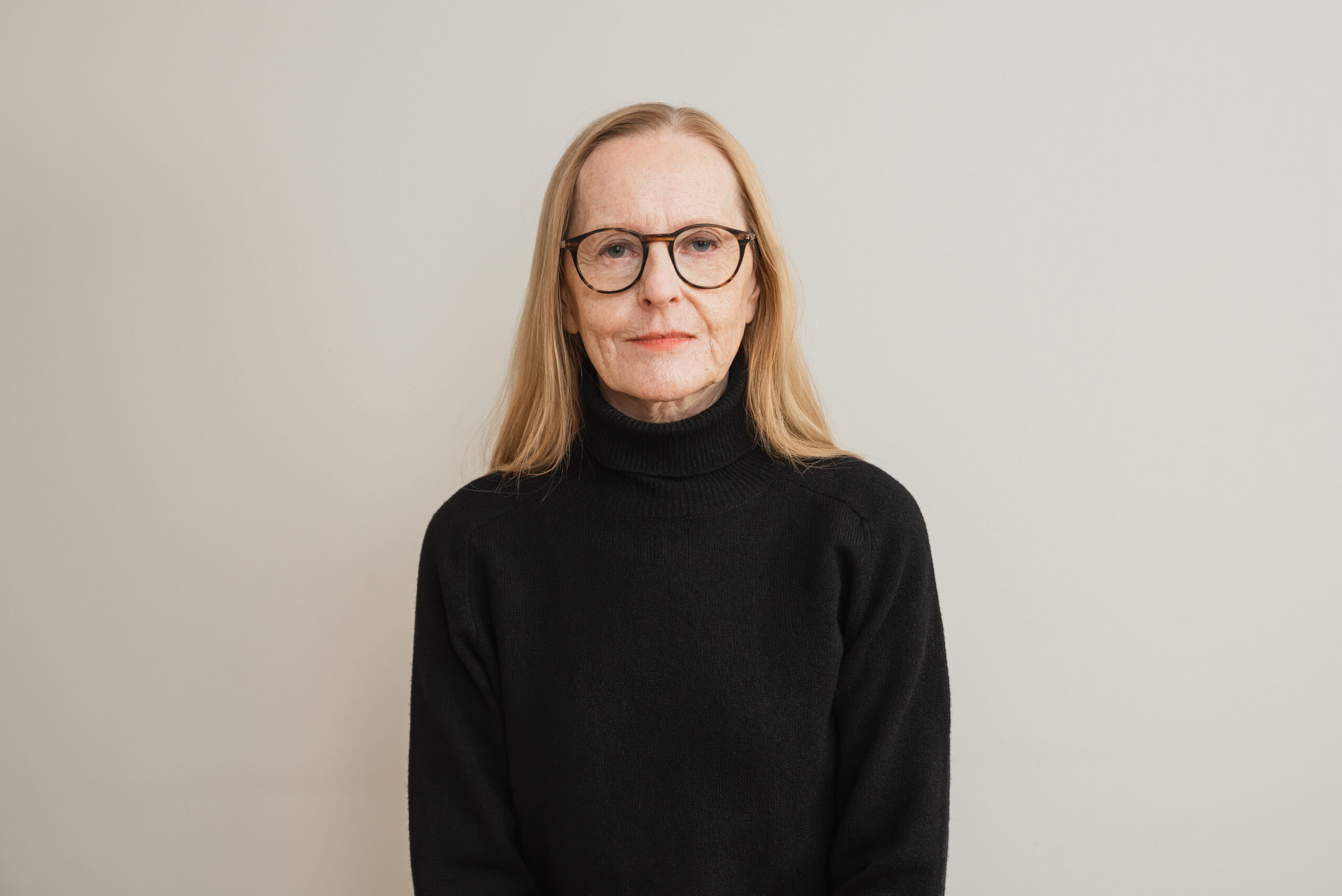 En person med långt blont hår och mörk polotröja står framför en ljus bakgrund.